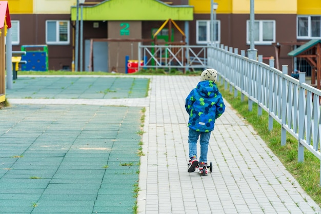 A child in a jacket and blue jeans rides a scooter on the sidewalk in the yard The concept of children's safety on the street A child without protective equipment is engaged in active recreation