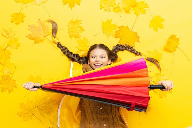 The child lies on the autumn maple leaves and holds out a red and pink umbrella smiling broadly It's autumn time for a happy little girl in a yellow raincoat and sweater