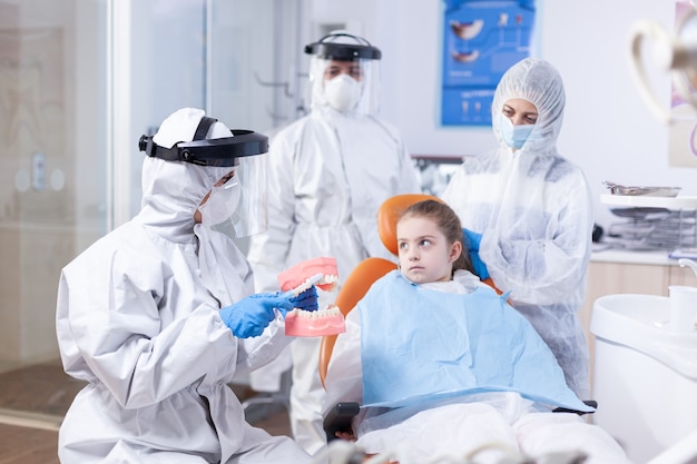 Child looking scared at parent sitting in dental chair. Little girl in ppe suit during covid19 and mother listening stomatolog talking about tooth hygine in dentistiry clinic holding jaw model.