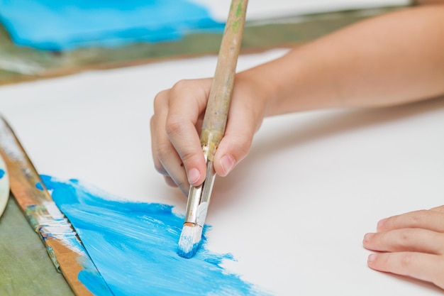 Child paints a picture by blue gouache Kid drawing at white paper The hand and paint brush Closeup selective focus