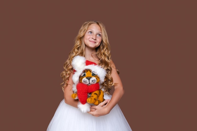 Child plays with cute little Christmas tiger toy in white dress on brown