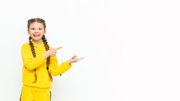 The child points to your advertisement A little girl with two pigtails in a yellow sports suit on a white isolated background