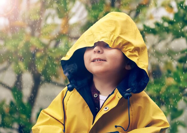 Photo child rain and raincoat protection in winter for fashion jacket or cold weather outdoor little boy male toddler and kid in drizzle by tree bokeh or environment with windbreaker for chilly season