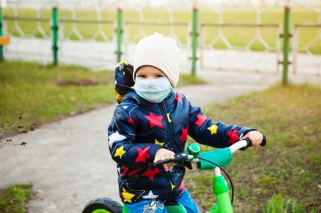 Child on a ride in a city park in a medical mask rides a bicycle. Walking on the street during the quarantine period of the coronavirus pandemic in the world. Precautions and teaching children.