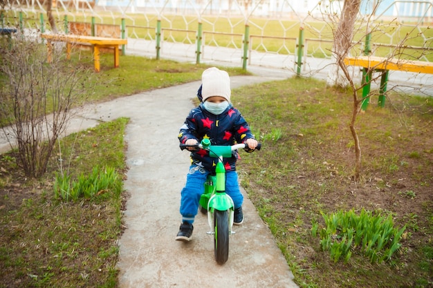 Child on a ride in a city park in a medical mask rides a bicycle. Walking on the street during the quarantine period of the coronavirus pandemic in the world. Precautions and teaching children.