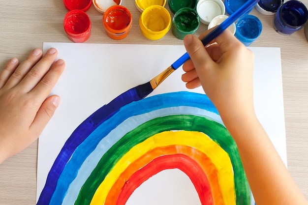 A child's hand draws a rainbow with a brushMany cans of red yellow green color on the mirror table in the classroomFemale fingers hold open blue paintThe concept of school art education