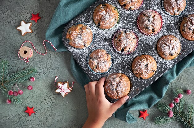 Child's hand taking a blueberry muffin with sugar icing from a baking tray with Christmas decorations