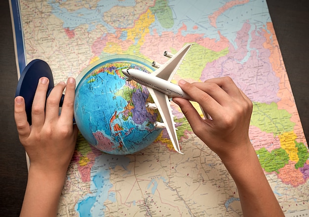 Child's hands are holding a globe and an airplane. World map background. Travel concept.