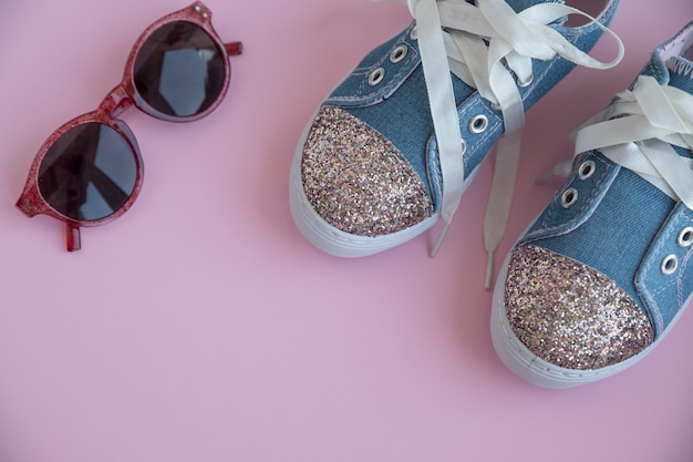 Photo child's textile lace sneakers. girls shoes on pink wall.fashion kid's footwear.smart casual fashionable denim and shiny shoes.sunglasses and trendy kids sports shoes.copy space. selective focus