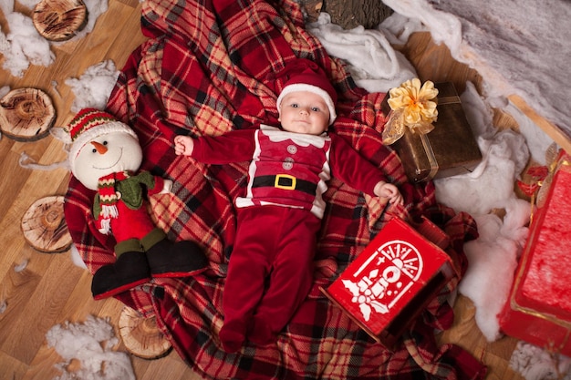 A child in Santa Claus costume rests on a red plaid surrounded by toys