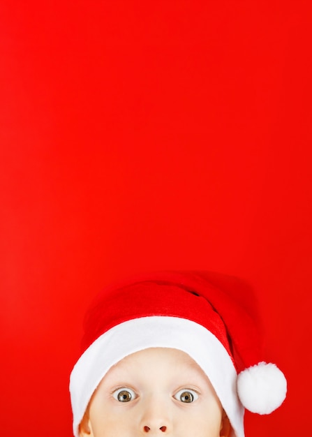 Child in a Santa hat on a red background looks out in surprise from the bottom of the frame