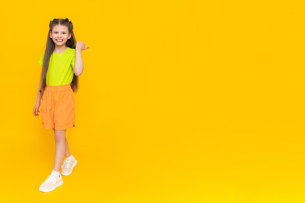 A child in shorts and a Tshirt Happy childhood A little girl with long hair enjoys the summer warmth inside and points to your advertisement on a yellow isolated background Copy space Banner