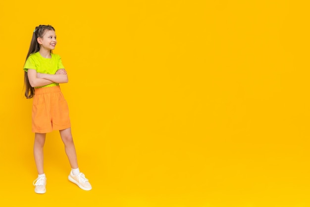 A child in shorts and a Tshirt Happy childhood A little girl with long hair enjoys the summer warmth inside and points to your advertisement on a yellow isolated background Copy space Banner