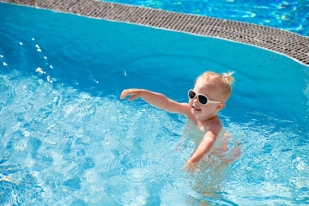 The child splashes in the pool with clear water in the summer illuminated by the sun.