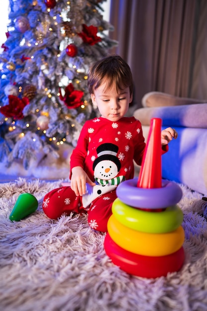 Child stacks colorful toys by the festive Christmas tree A young child in festive pajamas assembles colorful toys next to a Christmas tree adorned with lights and ornaments