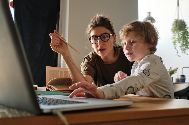 Child using laptop to do his online homework together with teacher at table