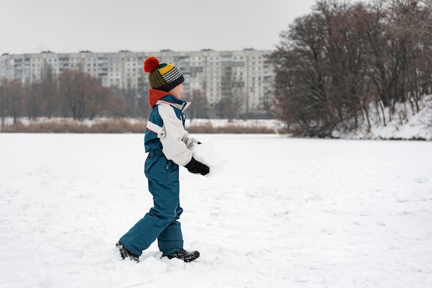 Child walks through snow-kept field. Boy in warm clothes plays outside in winter against the background of high-rise buildings.