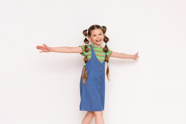 The child walks with his arms open in different directions in a summer denim sundress and smiles Beautiful little girl with long hair Copy space