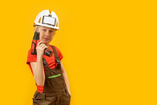 Child wears helmet and working overalls holds drill in his hands Boy 910 years old imitates builder yellow background Copy space