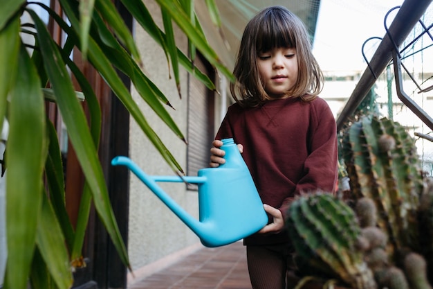 Photo child with brown hair holding blue watering can standing between plants at home balcony