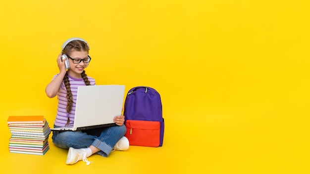 A child with a laptop and headphones A little girl is studying with a teacher online with books and a backpack Distance education for children Yellow isolated background Copy space