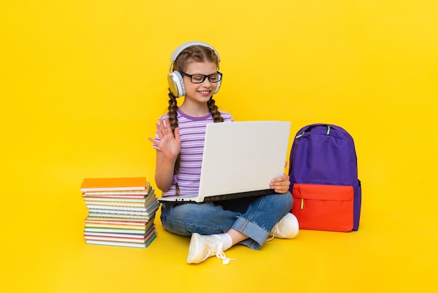 A child with a laptop and headphones A little girl is studying with a teacher online with books and a backpack Distance education for children Yellow isolated background