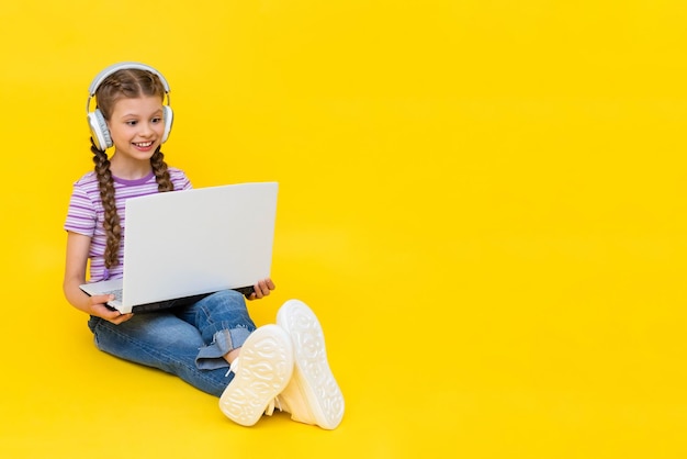 A child with a laptop A little girl sits on the floor and communicates online on a laptop wearing headphones Internet communication for children Yellow isolated background copy space