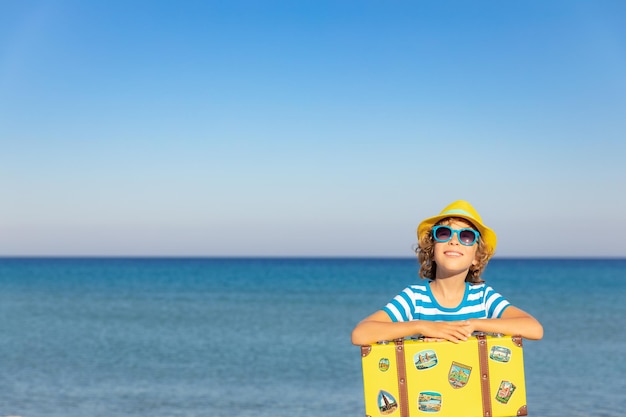 Child with vintage suitcase on summer vacation Girl having fun on the beach Kid sitting against sea and sky background  Travel and adventure concept