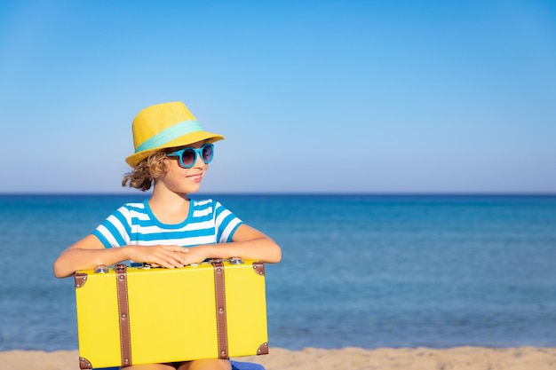 Child with vintage suitcase on summer vacation Girl having fun on the beach Kid sitting against sea and sky background Travel and adventure concept