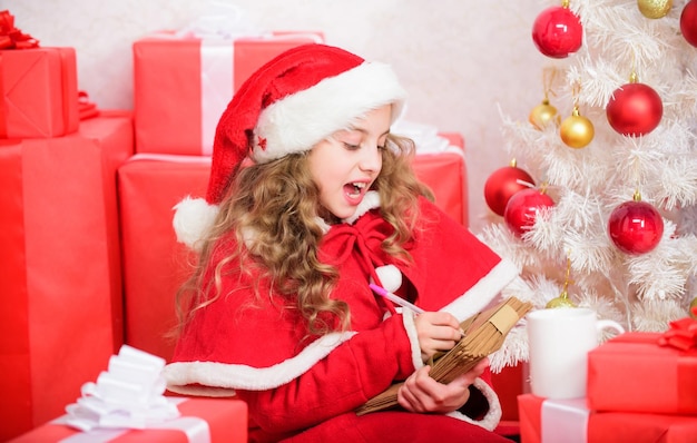 Child write letter to santa claus Believe in miracle Letter for santa Wish list Child santa costume enjoy christmas eve Girl little kid hold pen and paper near christmas tree writing letter