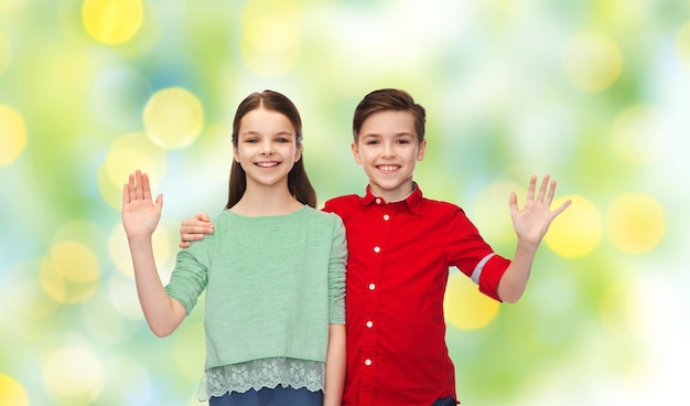 childhood, friendship, gesture and people concept - happy smiling boy and girl hugging and waving hand over green summer holidays lights background