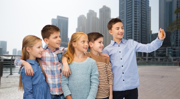 childhood, friendship, technology and people concept - group of happy children talking selfie by smartphone over dubai city street background