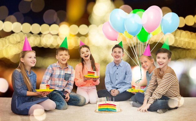 childhood holidays friendship and people concept happy smiling children in party hats with rainbow birthday cake and balloons over festive lights background