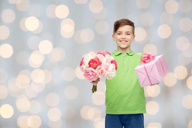 childhood, holidays, presents and people concept - happy boy holding flower bunch and gift box over lights background