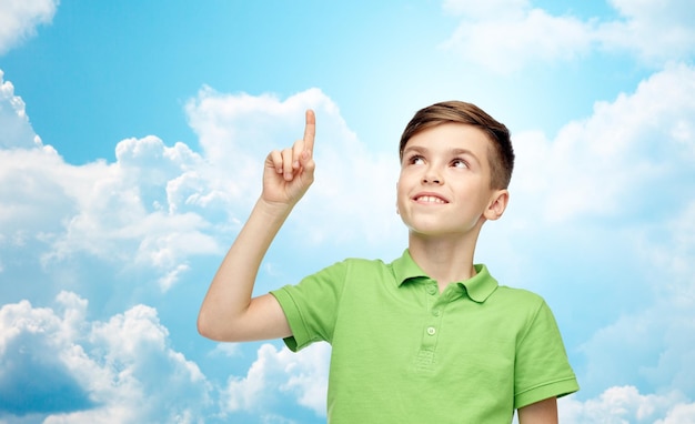 childhood, idea, inspiration and people concept - happy smiling boy in green polo t-shirt pointing finger up over blue sky and clouds background