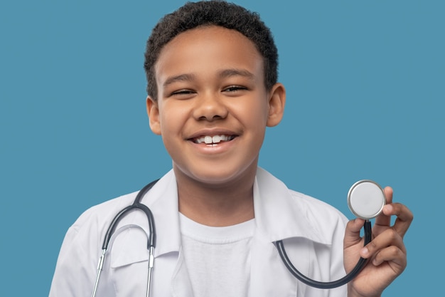 Childhood, joy. Happy dark skinned laughing boy in white coat showing medical stethoscope on blue background in studio