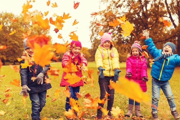 childhood, leisure, friendship and people concept - group of happy kids playing with autumn maple leaves and having fun in park