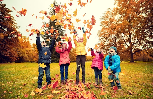 childhood, leisure, friendship and people concept - group of happy kids playing with autumn maple leaves and having fun in park