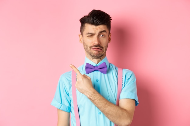 Childish young man acting sad and fake crying while pointing finger upper left corner, complaining on something, standing over pink background.