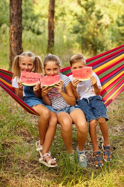 Children eat watermelon and joke outdoor sitting on a colorful hammock