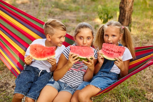 Children eat watermelon and joke outdoor sitting on a colorful hammock