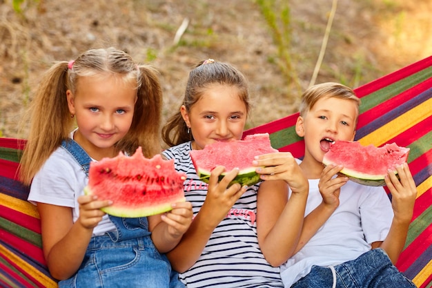 Children eat watermelon and joke outdoor sitting on a colorful hammock