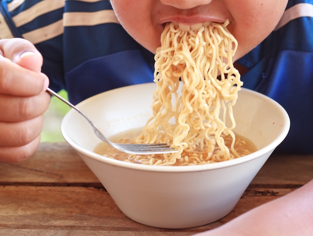 Children eating his instant noodle in white bowl on wood table,Focused on his mouth.