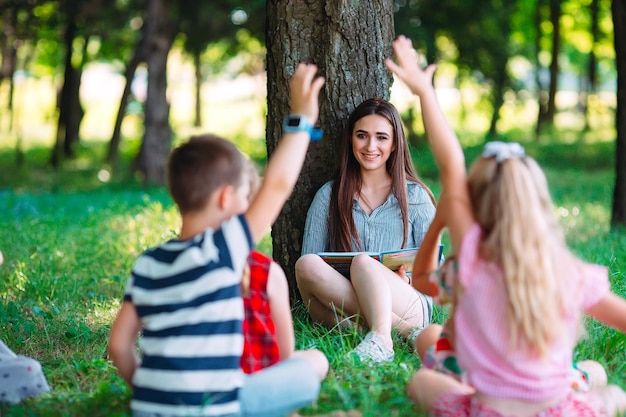 Children and education, young woman at work as educator reading book to boys and girls in park.