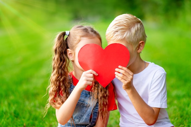 Children a girl and a blonde boy kiss behind a big red heart in summer on a lawn on green grass the concept of a valentine's day holiday space for text