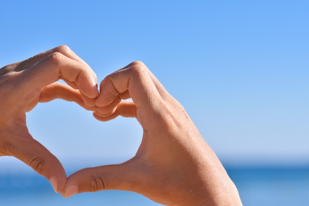 Children hands in the sand show a heart gesture against the blue sky and sea