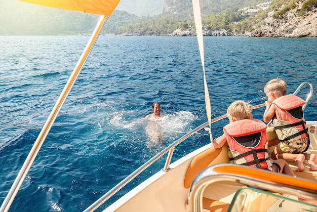 Children in life jackets sit on boat watching mother swimming in sea against coast of Marmaris