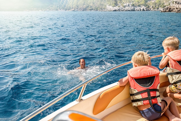 Children in life jackets sit on boat watching mother swimming in sea against coast of Marmaris