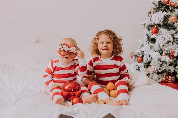 Children in red and white pajamas try on funny glasses with Santa Claus sitting in bed. lifestyle. brother and sister celebrating Christmas. boy and girl are playing at home. High quality photo