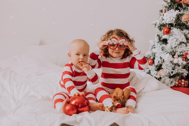 Children in red and white pajamas try on funny glasses with Santa Claus sitting in bed. lifestyle. brother and sister celebrating Christmas. boy and girl are playing at home. High quality photo